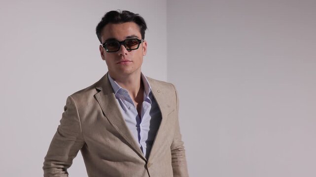 Serious man in beige suit and sunglasses standing with clasped hands, placing them in pockets, turning and walking away on white background.