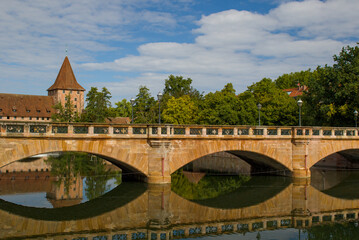 The ancient sandstone arches of the Max-Brucke span the Pegnitz River under a blue sky. Light warms historic walls as trees frame the skyline. The Bavarian landscape conveys the beauty of Nuremberg.