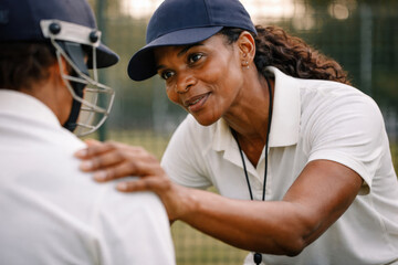 Supportive female cricket coach mentoring and encouraging young player with hand on shoulder during practice session on outdoor field