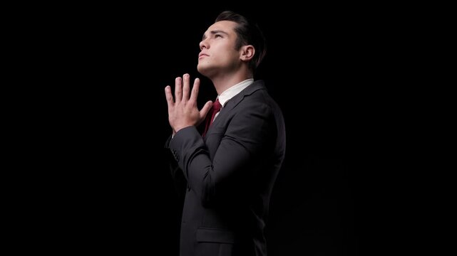 Side view of a stylish man praying with clasped hands and closed eyes, then looking up, turning to the camera, and walking away on a dark studio background.