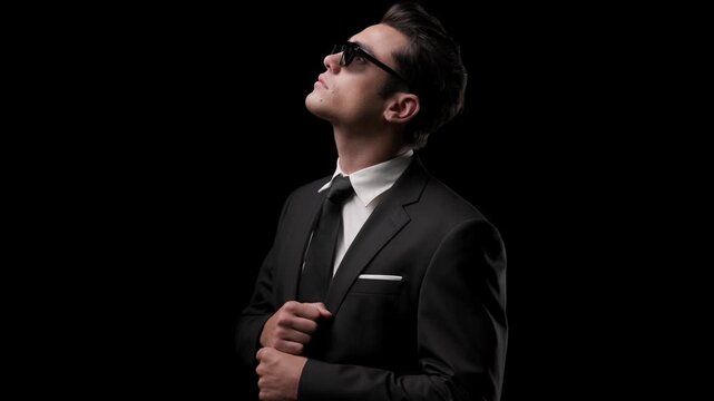 Stylish man in sunglasses adjusting suit jacket while looking up, then looking down and buttoning blazer on dark studio background.