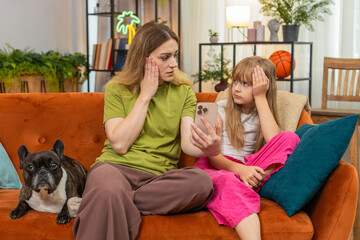 Mother and daughter lose game with French bulldog at home using smartphone shrug frown feeling upset. Woman, girl with dog on sofa react to fail game retry tap screen seek help support very sad mood