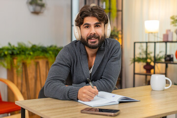 Indian man at home listens to audiobook with headphones takes notes in notebook stays focused....