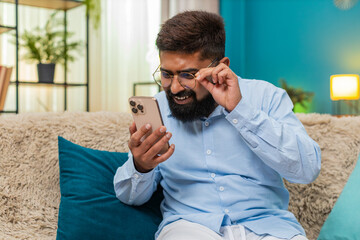 Indian man at home wearing glasses squints reading smartphone text holding screen closer focused....