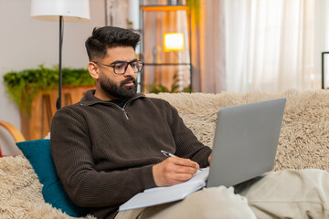 Indian man at home writes carefully in notebook while reading laptop screen, focused on learning....