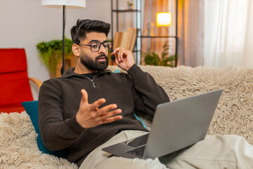 Indian man at home speaks on laptop video call using wireless headset, gesturing while talking with client. Hispanic guy on sofa works in call center remotely, staying focused and confident online