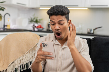 Indian man at home watches wow video on smartphone feels shock laughs smiles excited by news. Arabian guy on sofa shares link in messenger sends to friends writes short reply and posts reaction emoji
