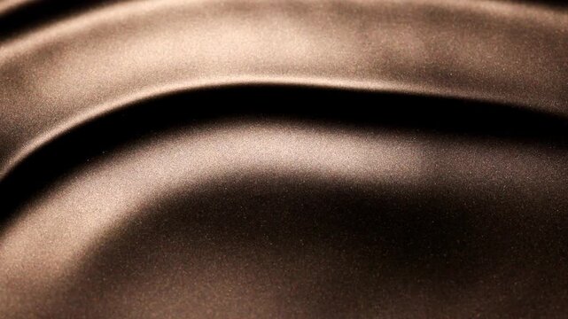 Abstract slow-motion video of large waves and deep ridges created by shifting dark golden liquid paint and dramatic variation of light and shadows.