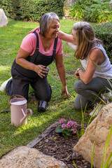 Diverse female friends kneeling in backyard garden planting flowering plant with watering can