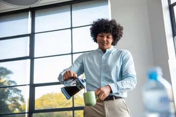 Man pouring coffee from glass pot into green mug by grid window in office break area