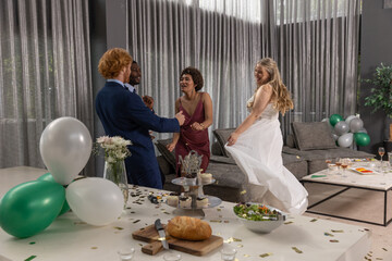 Obraz premium Diverse group of four in wedding attire dancing at home with confetti, balloons, champagne