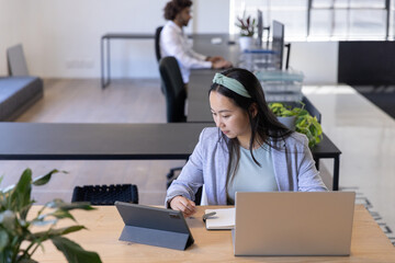 Diverse coworkers writing in notebook and typing on desktop, referencing tablet at office desk