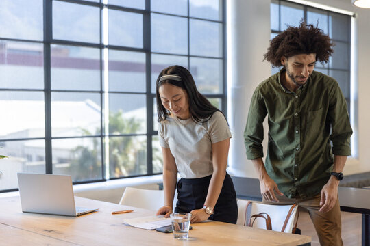 Diverse coworkers standing by conference table reviewing printed documents using laptop - Powered by Adobe
