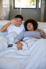 Fototapeta premium Diverse male partners shielding eyes and cuddling under duvet on bed in bedroom with smartphone