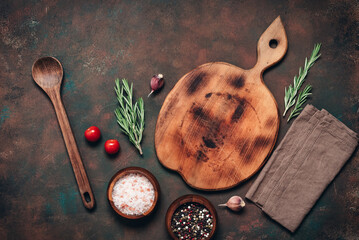 A culinary backdrop with an empty wooden cutting board, a wooden spoon, and various spices and herbs, dark grunge background. Top view, flat lay, copy space.