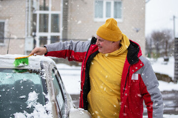 Man clearing snow from car hood in winter weather while wearing warm clothing in a residential area