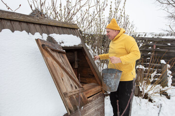 Man in yellow sweater collects water from wooden shed covered in snow during winter day in the backyard