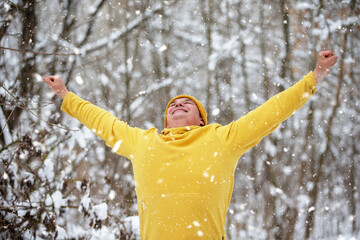 Man in yellow sweatshirt enjoying snowfall in winter forest, arms raised in celebration on a snowy day