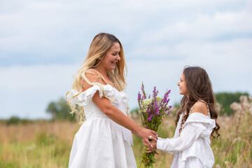 Mother and daughter joyfully share a moment in a field wearing white dresses while holding flowers
