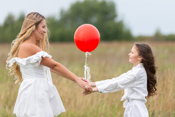 Woman and girl sharing a joyful moment with a red balloon in a grassy field during a cloudy day