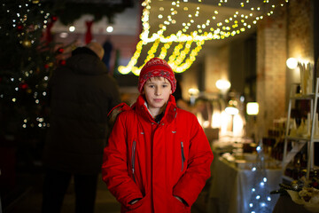 Child in red coat and hat stands at festive market with lights and holiday decorations in winter season