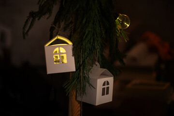 Decorative white houses hanging on a Christmas tree with warm lights in a cozy room