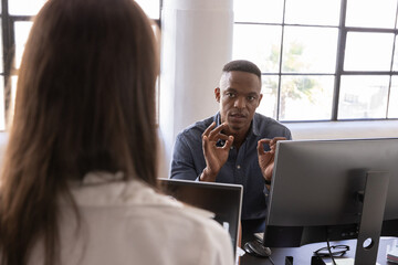 Diverse coworkers gesturing and discussing project at office desk with laptop, monitors