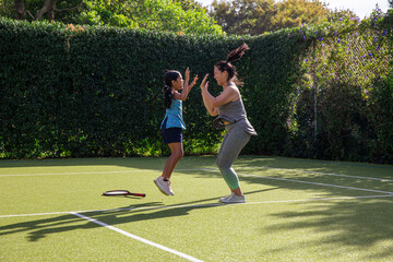 Obraz premium Diverse mom and daughter in gear jumping and high-fiving on tennis court with racket, white lines