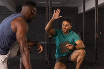 Diverse male trainer and client kneeling at gym squat rack guiding squat with tablet, copy space
