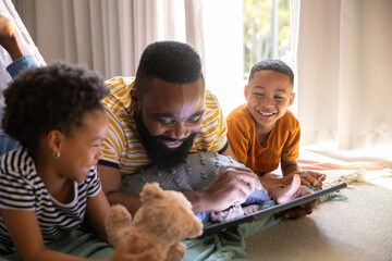 Diverse family lying on rug in living room using tablet computer with pillows, stuffed teddy bear