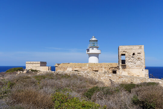The Sentinel of the Sea: A crisp white lighthouse stands tall against a deep blue sky, overlooking a rugged coastal landscape and sitting upon historic, sun-baked stone walls, symbolizing solitude and