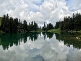 Obraz premium Calm mountain lake at Grantrisch in the Bernese Oberland, Switzerland, featuring a unique mirror-like reflection of dramatic cloud formations and surrounding dark fir trees