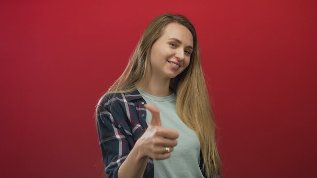 Woman smiling against a bold red background, giving a thumbs up, dressed casually in a plaid shirt and grey t-shirt, conveying positivity and confidence.