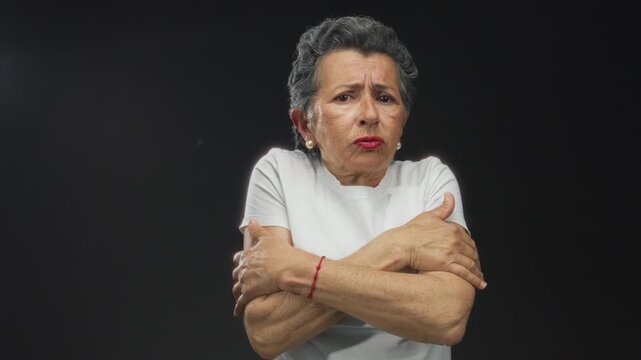 Senior woman with grey hair looking cold against a black background, clutching her arms and wearing a white shirt, expressing chilly discomfort.