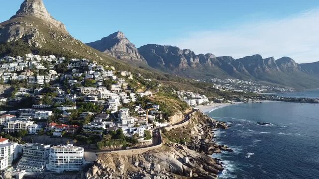 Cinematic drone footage of the picturesque Camps Bay coastline and Victoria Road winding between the ocean and mountains; aerial shot of the famous white sand beach and luxury apartments in Cape Town