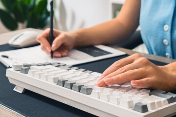 A student is sitting at a desk, typing on a keyboard with one hand and writing in a notebook with a pen in the other. Learning. Close-up.