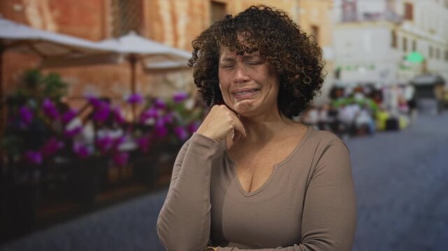 Hispanic woman covering face with hand at outdoor restaurant terrace beside flower pots and tables; sadness.