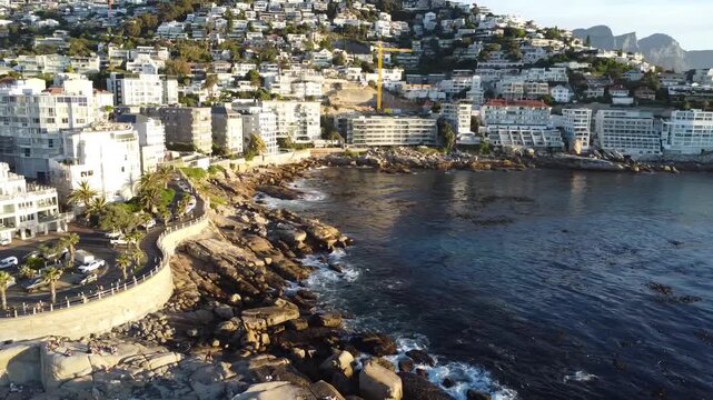Aerial drone view of the scenic Victoria Road winding along the coast of Camps Bay Cape Town; luxury apartments overlooking the white sand beach and turquoise Atlantic Ocean in South Africa.