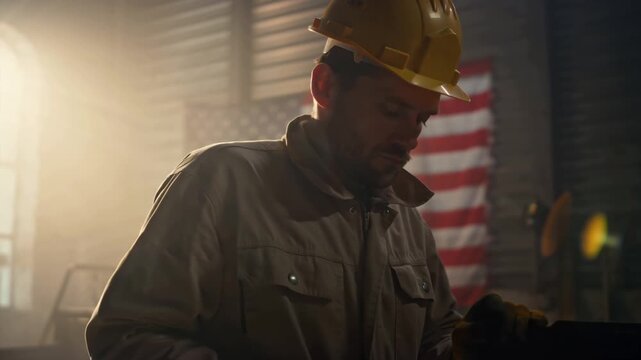 Cinematic 4K serious American male worker, wearing rugged work uniform hard hat, performing tasks sunrise in an industrial setting. The American flag waves in the background. Warm golden sunlight