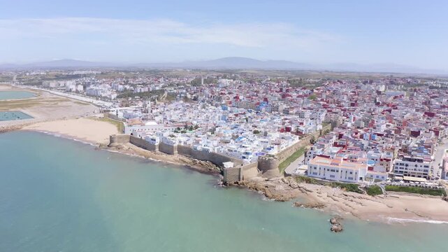 Assilah, Morocco - May 8, 2024: Wide orbit drone shot of Assilah, Morocco, featuring the historic city surrounded by ancient walls, overlooking the Atlantic Ocean with mountains in the background, hig