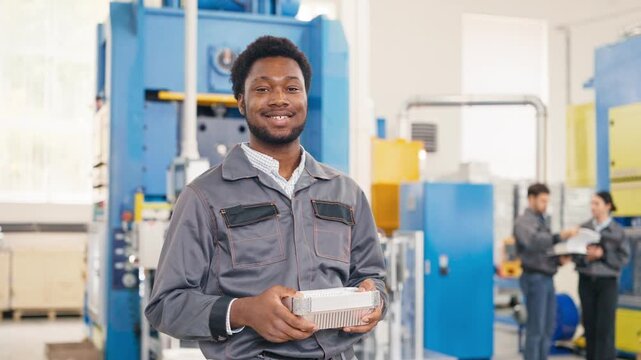 Smiling African American male standing in production hall holding stack of foil containers. Confident expression showing satisfaction with work results. Colleagues in background discussing production.