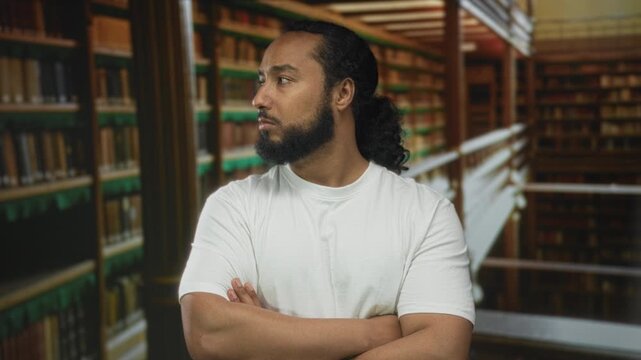 Man in white t shirt with arms crossed beside tall bookshelves in a building, looking sideways with beard and tied hair; thoughtful study.