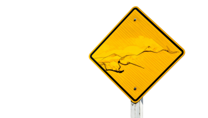 Damaged, yellow diamond road sign warns of cracks, possibly a fissure. Against black