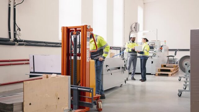 Male factory worker in safety jacket and helmet operating orange pallet lifter near machinery. In background colleagues in discussion using laptop. Industrial workspace with equipment and materials.