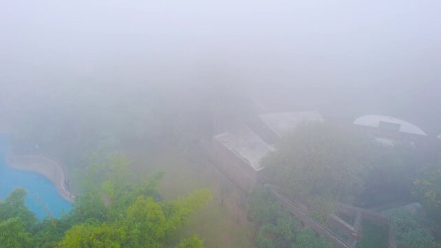 aerial drone shot in dense fog smog air pollution flying over dense green trees, blue pool and multiple brick red stone buildings showing the situation on small cities near delhi, gurgaon, noida