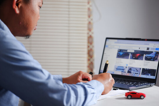 Buying and selling cars online. A man wants to buy a used car. He browses various online used car listings on his laptop to compare prices. He then notes it down.