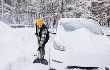 Man shoveling snow next to his electric vehicle, preparing for winter travel and dealing with...