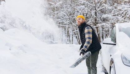 Young man shoveling snow after winter blizzard, clearing path around his car in cold weather