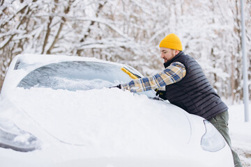 Man clearing snow and ice from car windshield using brush in winter preparing for daily commute