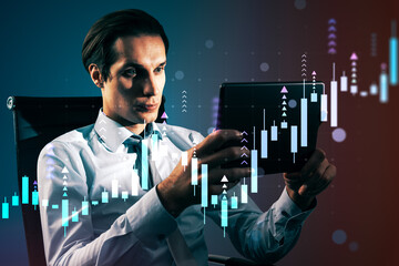 Businessman analyzing tablet with digital candlestick chart overlay in modern office environment under blue and red lighting tones.